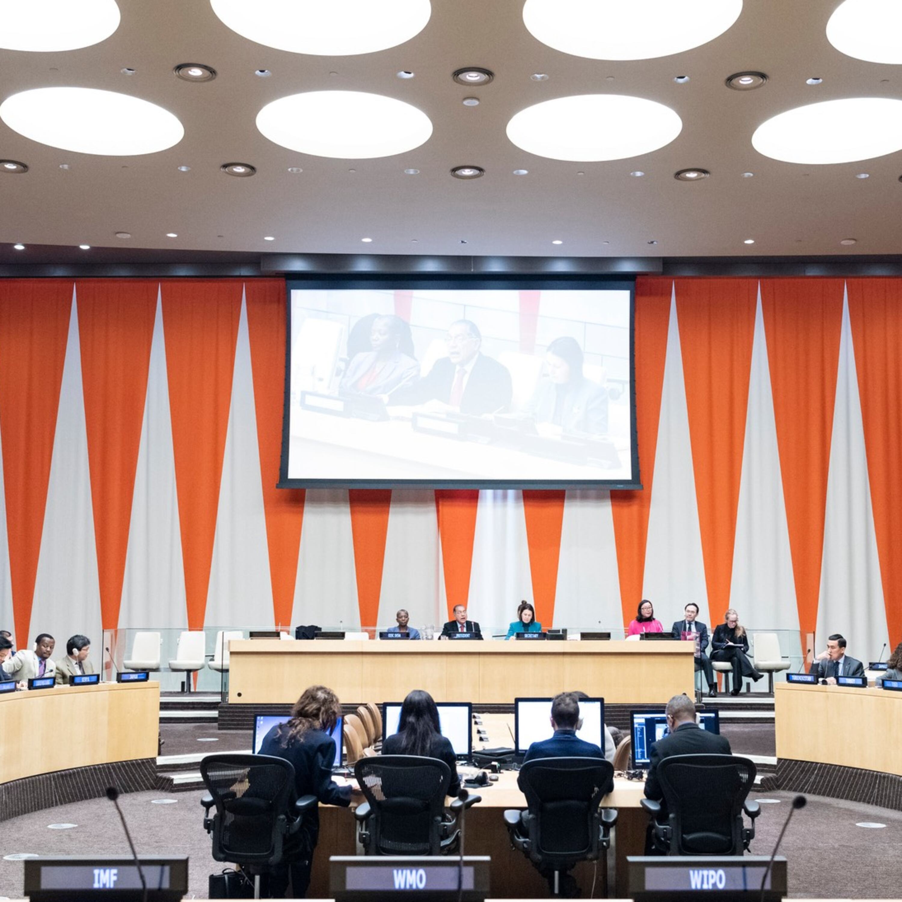 Wide view of the United Nations Economic and Social Council (ECOSOC) chamber during a formal meeting with delegates seated at desks with international organization placards such as World Bank, IMF, WHO, WIPO, and IFAD.