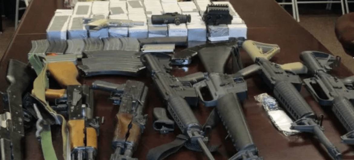 A collection of seized illegal weapons and ammunition laid out on a table, including assault rifles, handguns, and boxes of ammunition, in the context of gang violence in Haiti.