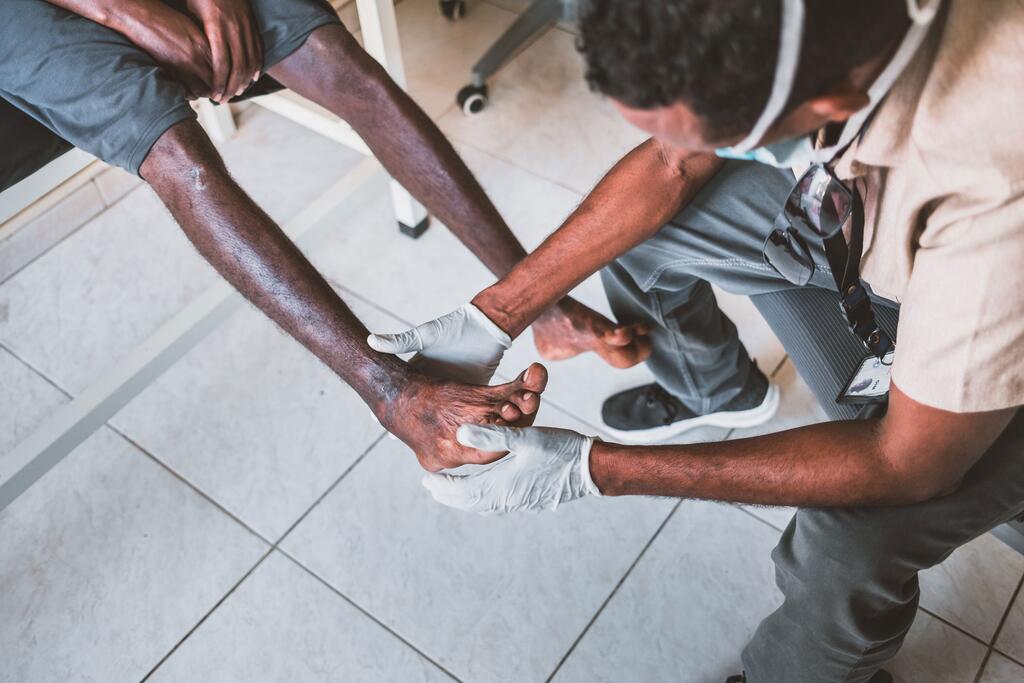 A medical worker wearing gloves examines the injured foot of a seated African migrant in a clinic setting.