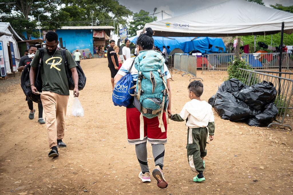 A migrant family, including a woman with a large backpack and a small child, walks through a temporary shelter area at the Lajas Blancas reception centre in Darien, receiving aid from IOM staff.