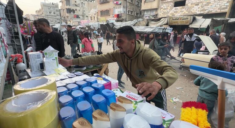 A Palestinian man sells household goods like cleaning supplies and tape at a makeshift street market in Khan Younis, Gaza. In the background, damaged buildings are visible, illustrating the 'survival economy' where educated youth take on simple jobs due to the ongoing conflict.