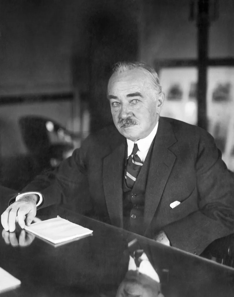 Elderly man in a suit sits at a polished desk with papers in a dim office.