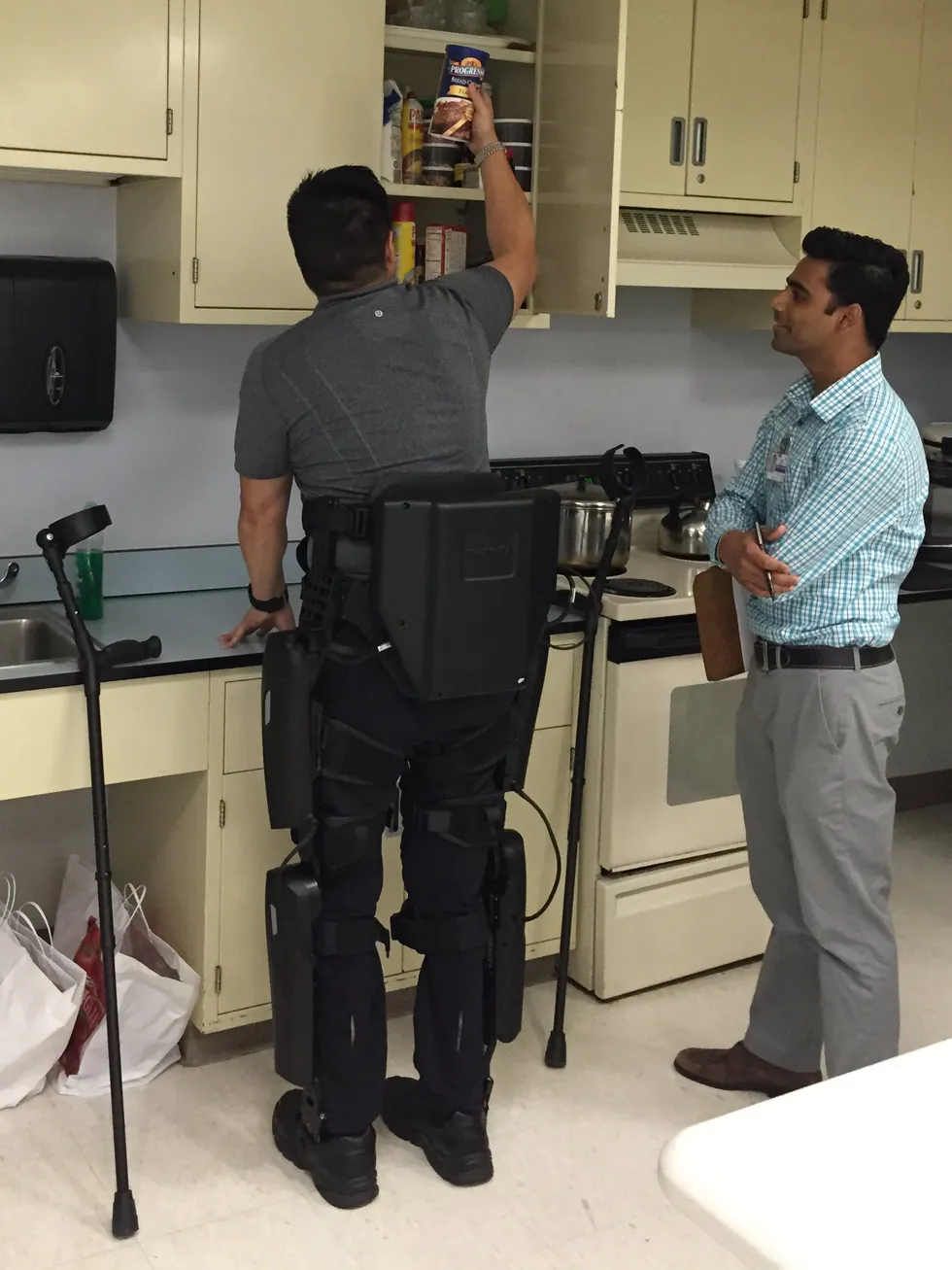 Man in a leg exoskeleton reaches into a kitchen cabinet while another observes.