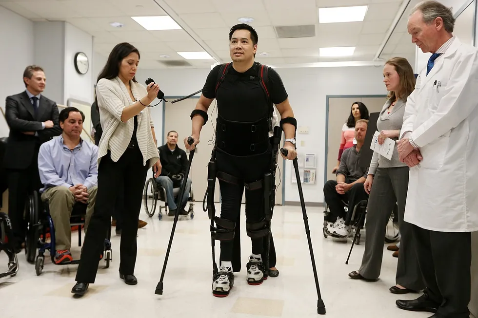 Man in robotic exoskeleton walks with canes during rehab demo as clinicians observe