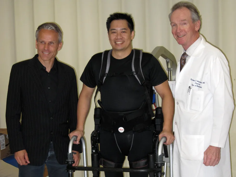 Patient in exoskeleton uses walker, flanked by doctor in lab coat and man in suit