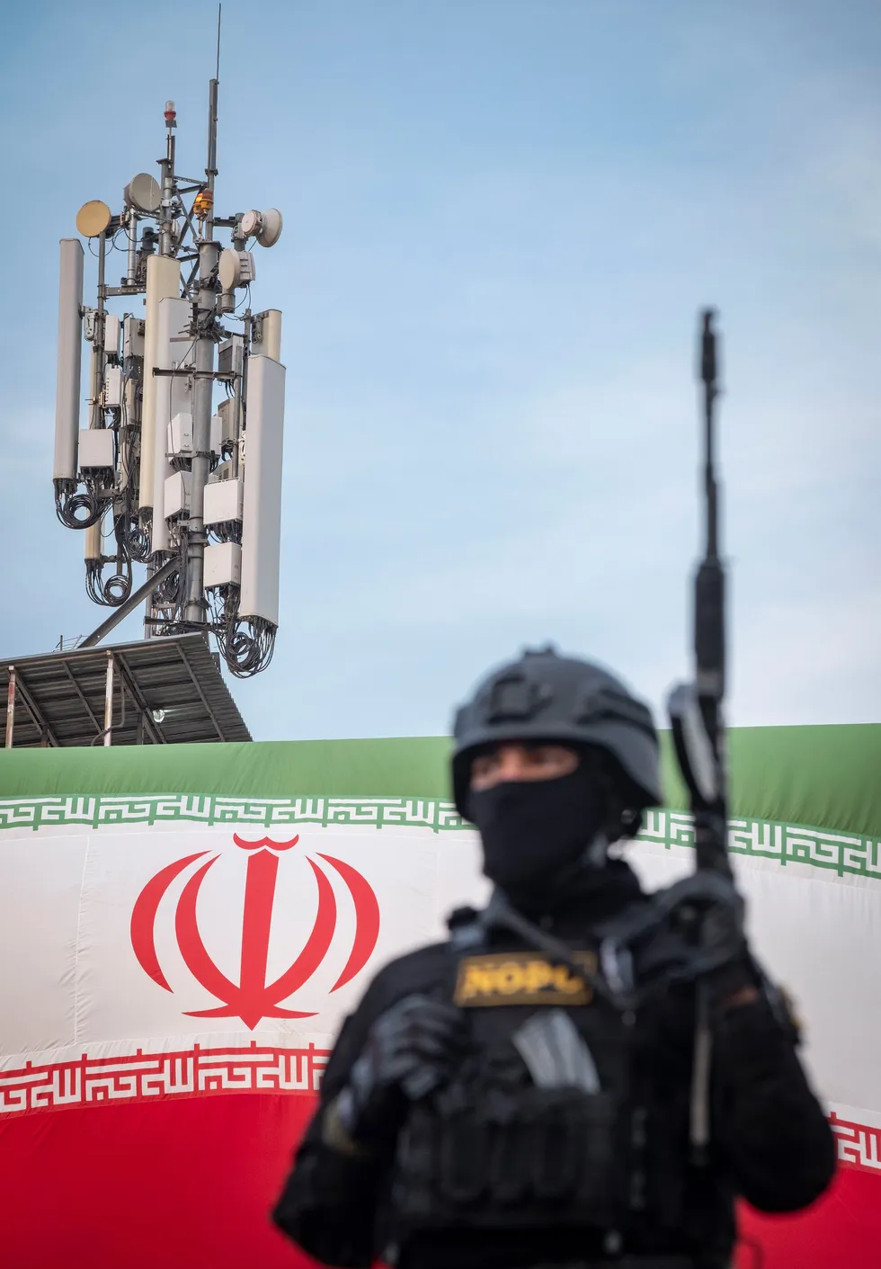 Photo of a helmeted soldier with a machine gun standing in front of an Iranian flag and cell tower.