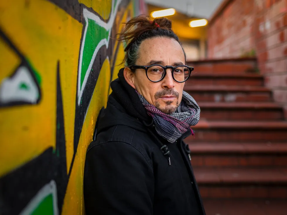 Man dressed in a coat and scarf leaning against a graffitied wall