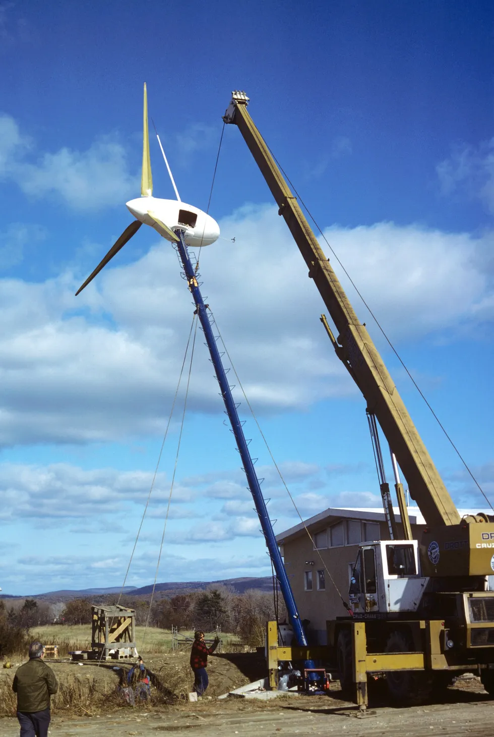 A crane in the midst of vertically erecting a wind turbine on a single pole    