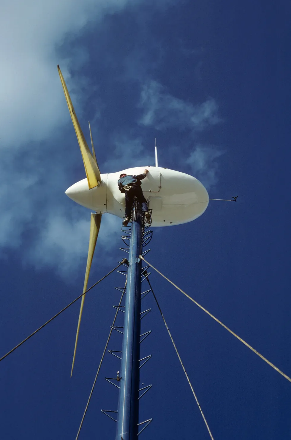 A man in a harness standing at the top of a wind turbine on a single pole, high in the air