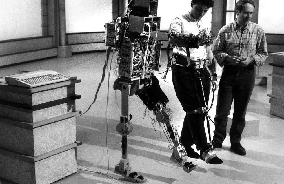 Black and white photo of a two-legged humanoid robot with its left leg raised, next to a man with his right leg raised while another man looks on.