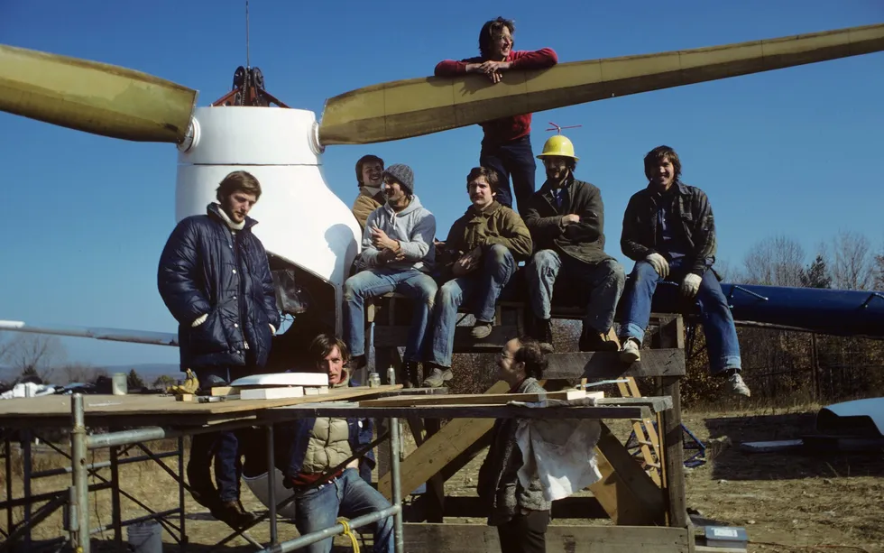 Nine men standing and sitting on scaffolding that holds up the rotor and blades of a wind turbine