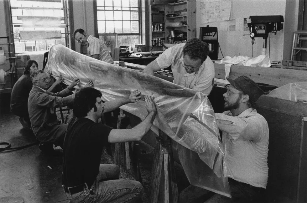 Six men squat around a turbine blade that\u2019s wrapped in plastic