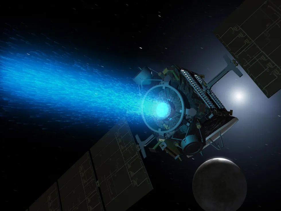 A boxy spacecraft with large solar cells flies through space, propelled by a blue exhaust from a thruster. 
