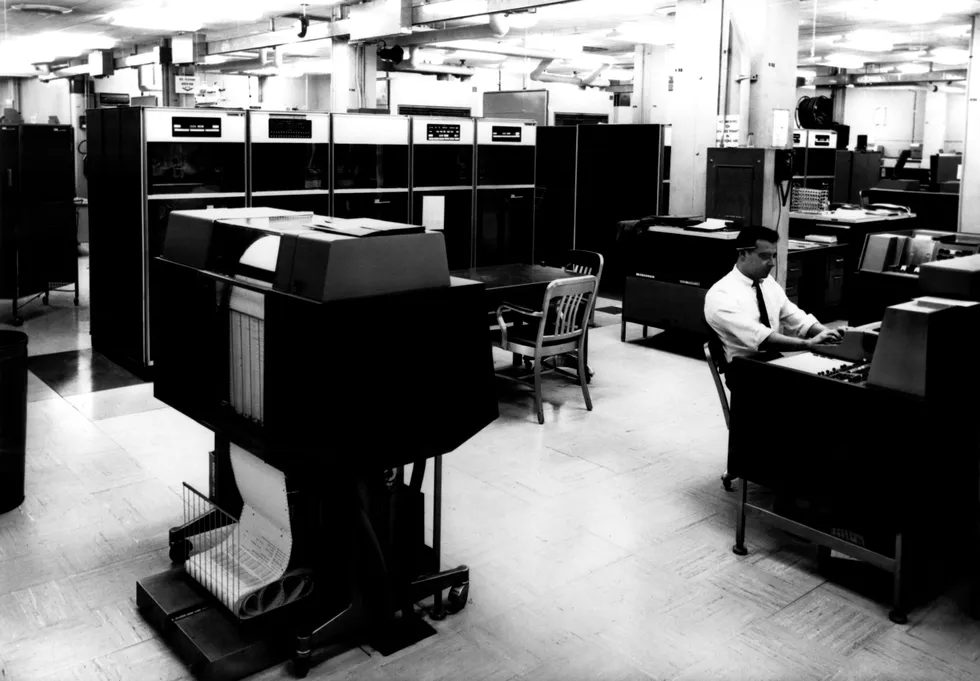 a computer center with printers and large cabinets and a man sitting at a computer keyboard