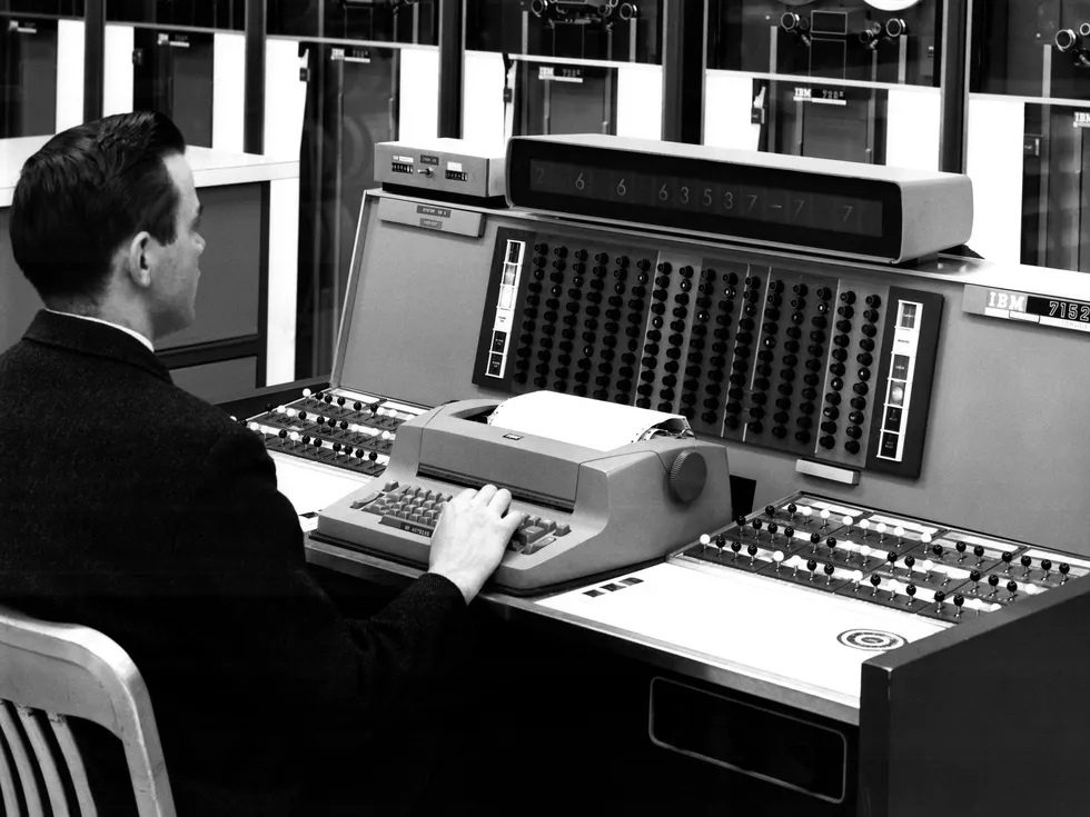a man at typewriter typing on keyboard with computer equipment in background; in the center