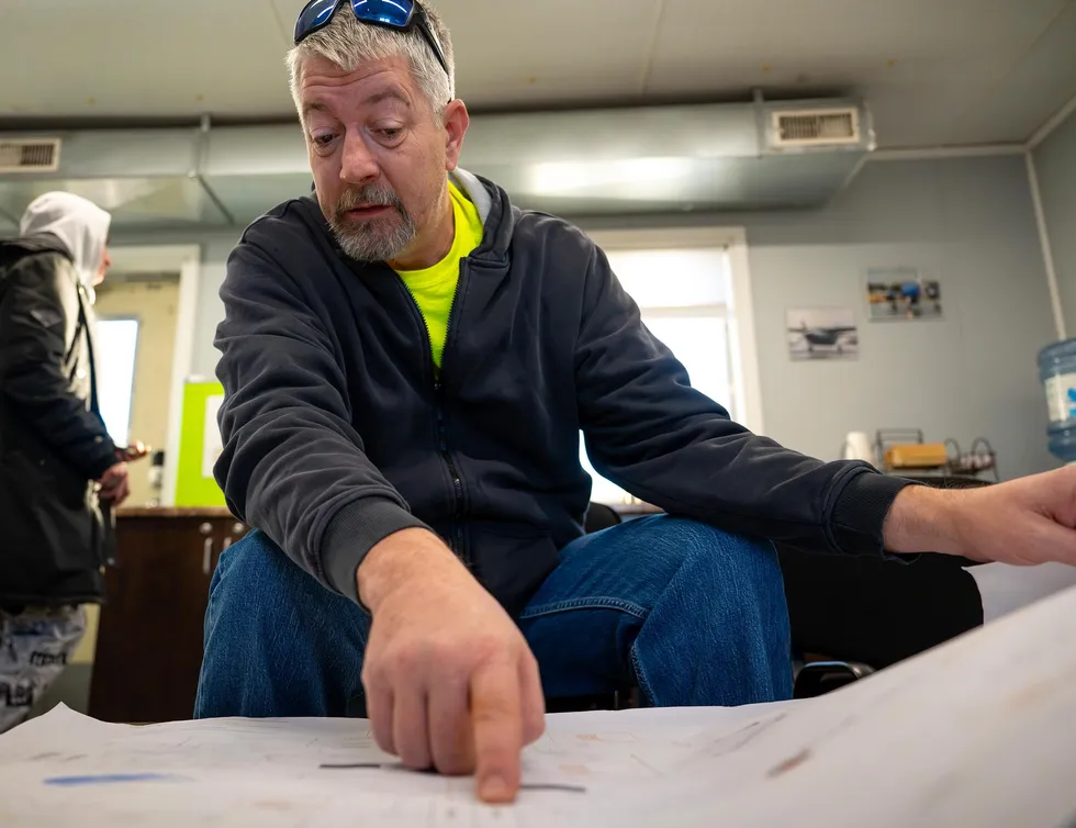 A man in an office points to something on a large piece of paper spread before him.  