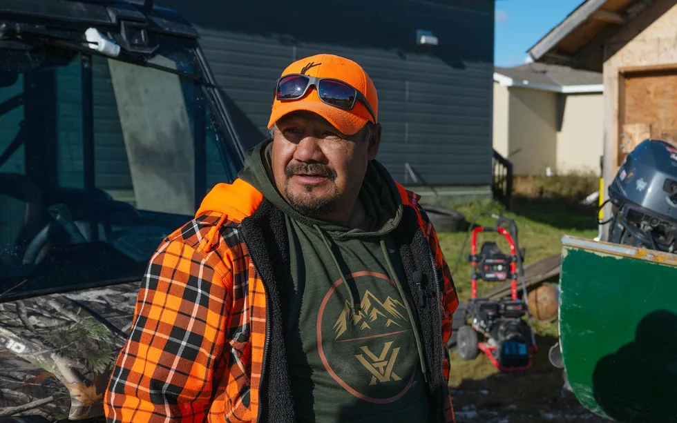 A man in an orange baseball hat stands next to a boat with an outboard motor in front of a house. 