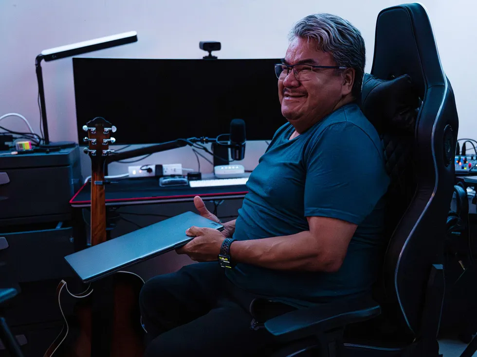 A photo shows a man holding a laptop and sitting in an office in front of a large computer monitor. 