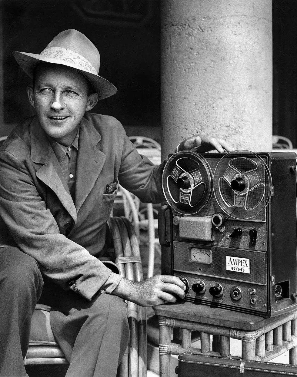 Black and white photo of a smiling man in a suit and hat sitting next to a reel to reel tape recorder.