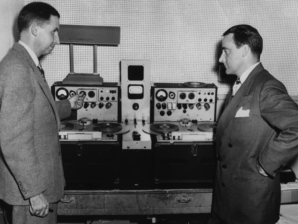 Black and white photo of two men in suits facing each other and standing in front of radio equipment. 