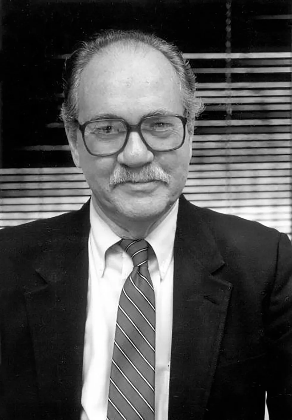 Headshot of man with moustache and glasses, in a business suit.