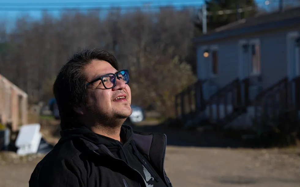 Person in glasses looks up, smiling, outside on a sunny day near small houses.