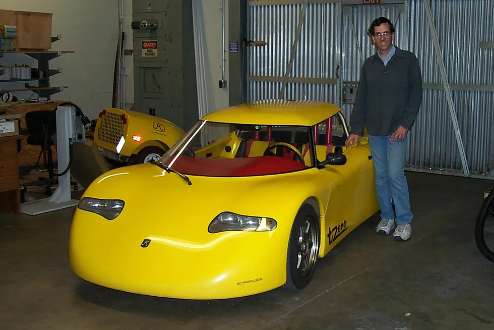 A man wearing white sneakers and blue jeans stands next to a yellow roadster with a bright red dashboard.