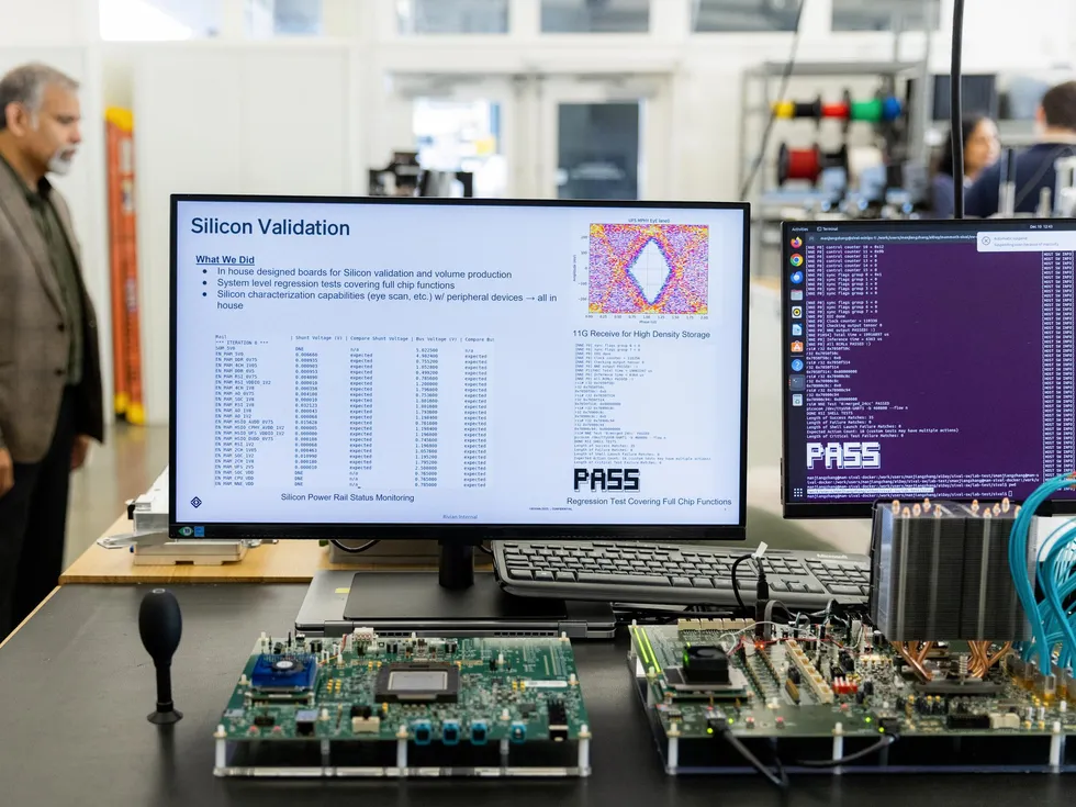 A man with a goatee wearing a blazer stands next to a bench with printed circuit boards and computer monitors.
