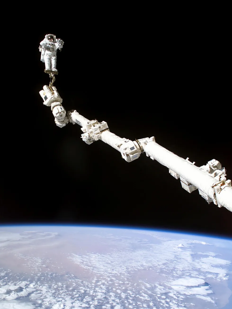 An Astronaut with their foot anchored to an extended robotic arm in low Earth orbit.