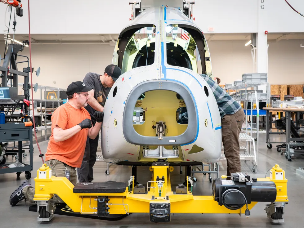 Color photo of three men building the body of a small airplane that\u2019s sitting on a yellow platform.